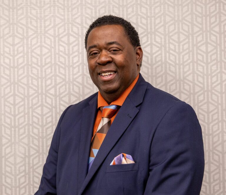 Professional portrait of Bruce White smiling in a brightly lit indoor setting.