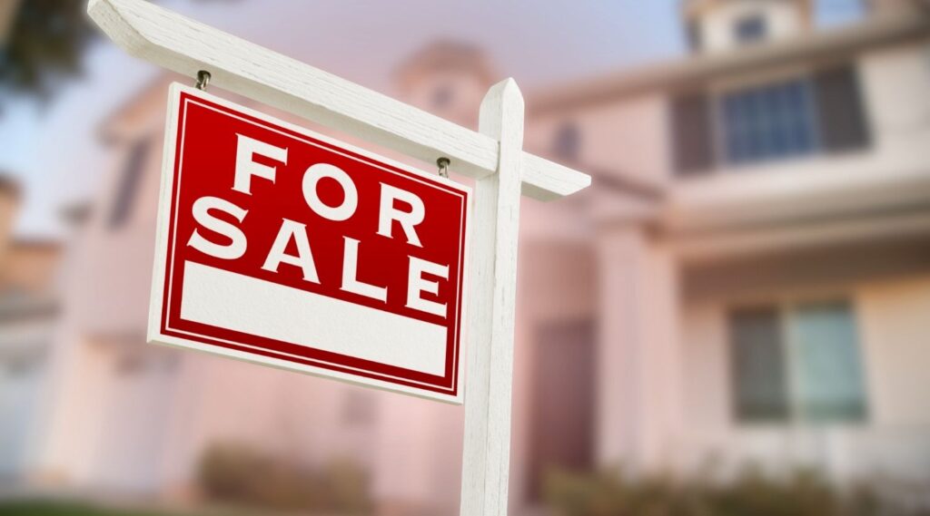 A red and white "For Sale" real estate sign posted in front of a blurred suburban house.