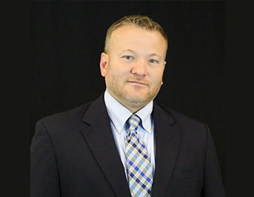 A professional portrait of a man with short brown hair and a beard wearing a navy blazer, light blue shirt, and a blue and grey plaid tie against a black background.