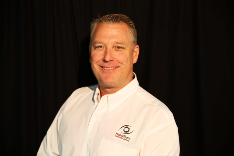 A smiling man with short hair wears a white button-down shirt featuring the HomeTeam Inspection Service logo, set against a solid black background.
