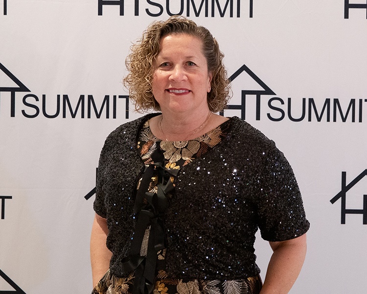 A woman with curly brown hair smiling in front of a white backdrop with SUMMIT logos, wearing a black sequined shrug over a gold and black floral dress.