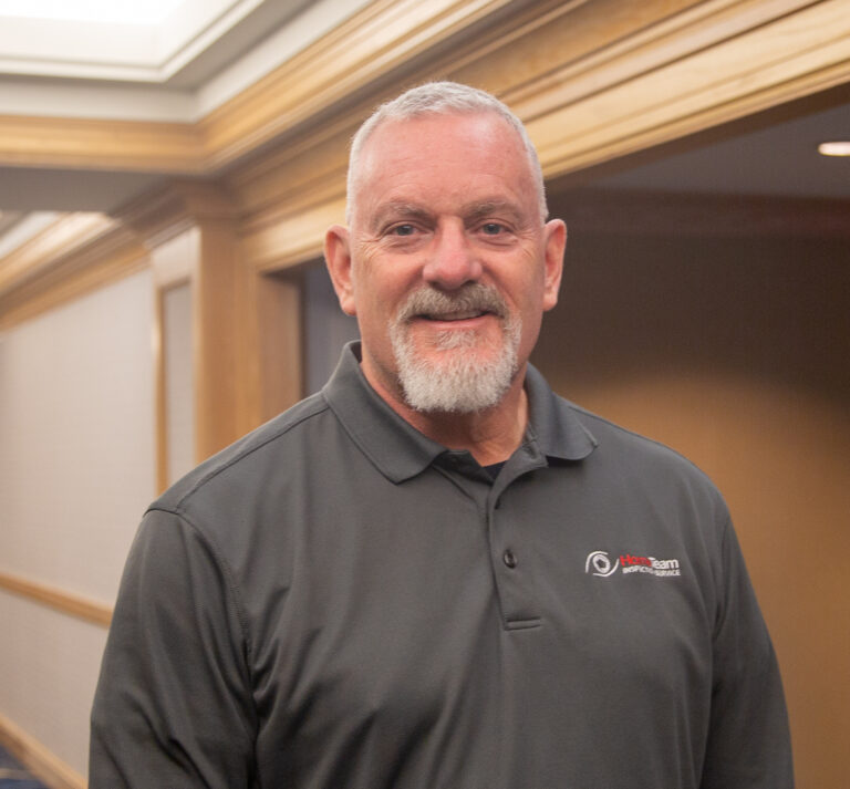 A headshot of a smiling man with short grey hair and a goatee, wearing a grey polo shirt with the HomeTeam Inspection Service logo.
