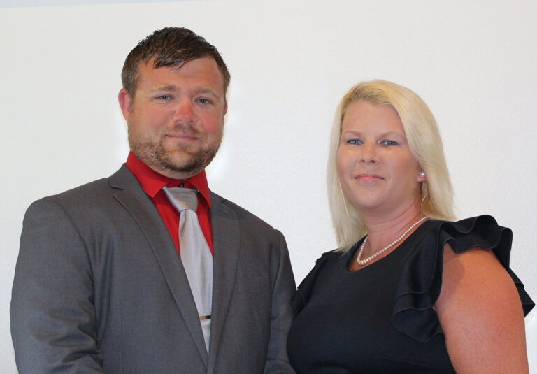 A portrait of Jason and Jamie Kulinski standing side-by-side against a plain white background; Jason is wearing a grey suit with a red shirt and silver tie, and Jamie is wearing a black dress with ruffled sleeves and a pearl necklace.