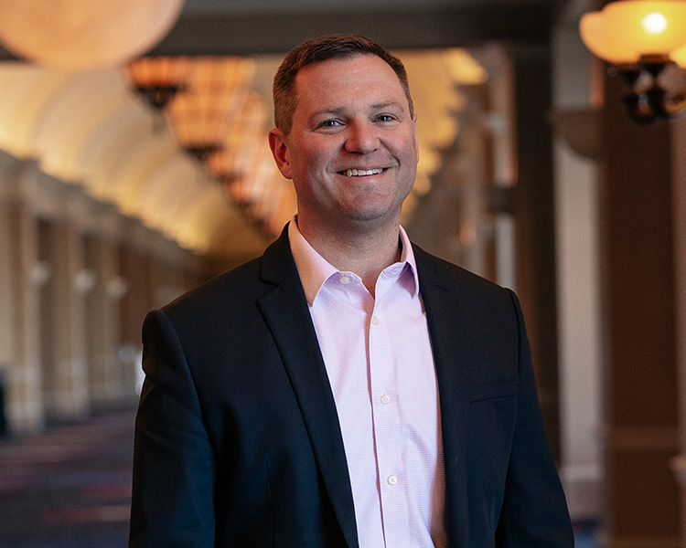 A professional portrait of a smiling man wearing a dark blazer and a light pink dress shirt, standing in a warmly lit, blurred hallway.