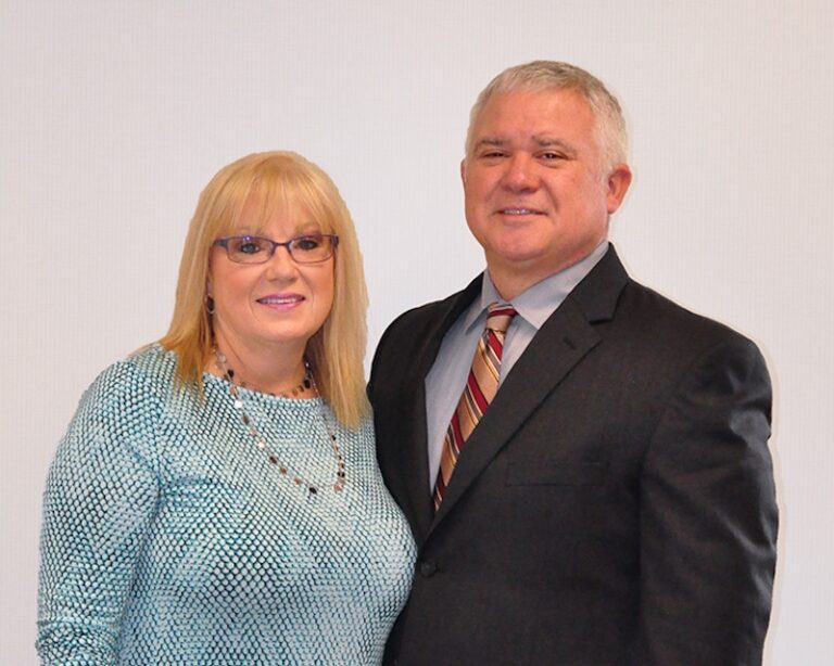 A professional portrait of a blonde woman in a light blue patterned top and a man with short grey hair in a dark suit and striped tie, both smiling against a light background.