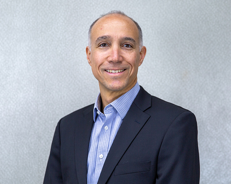 Professional headshot of Roberto Haza, smiling and wearing a dark blazer over a checkered blue button-down shirt against a neutral gray background