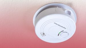 A white carbon monoxide alarm mounted on a textured ceiling with a red gradient background.