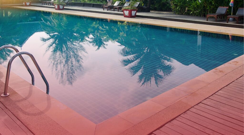 A clean, blue-tiled swimming pool reflecting palm trees, featuring a stainless steel ladder in the foreground and lounge chairs on the surrounding deck.