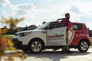 A man in a red shirt steps out of a white and red branded SUV featuring the "HomeTeam Inspection Service" logo on the open driver-side door.