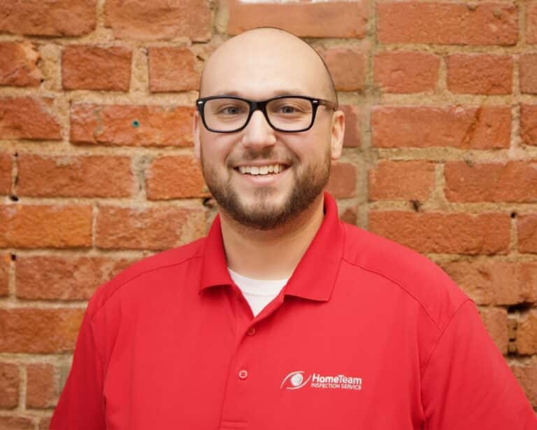 Headshot of smiling Adam Cane with glasses and a beard wearing a red HomeTeam Inspection Service polo shirt against an exposed red brick wall.