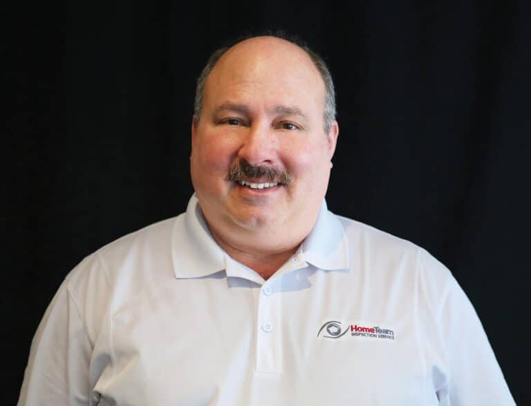 Bob Day, owner of HomeTeam of Minneapolis, is a smiling man with a mustache wearing a white HomeTeam Inspection Service polo shirt against a plain black background.