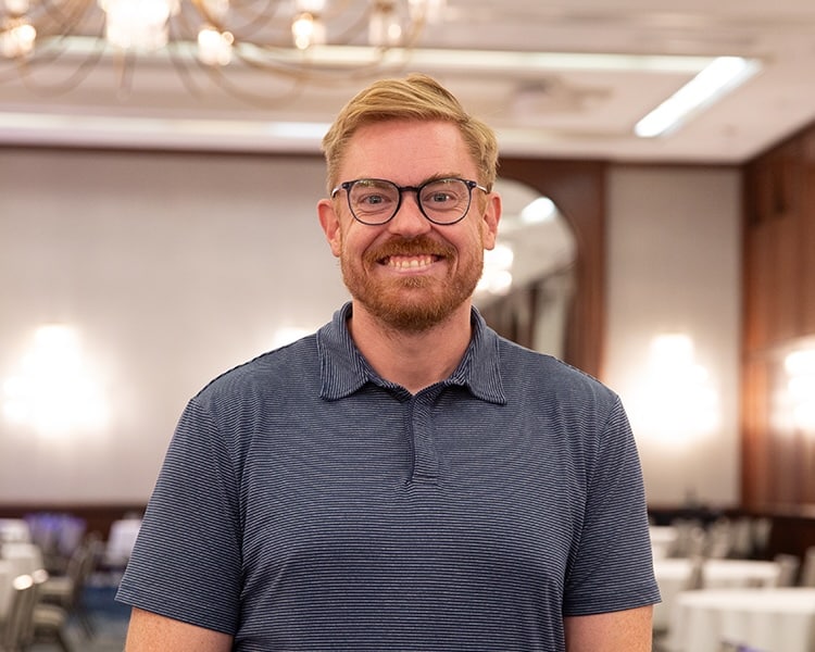 A smiling man with a beard and glasses wears a blue striped polo shirt while standing in a banquet hall with tables blurred in the background.