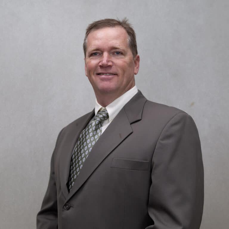Professional headshot of Jason Adams, a smiling man in a brown suit jacket and patterned tie, posing against a neutral gray background.