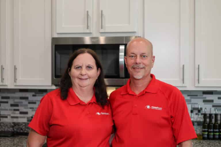 A man and a woman wearing matching red "HomeTeam Inspection Service" polo shirts smile while posing together in a kitchen.