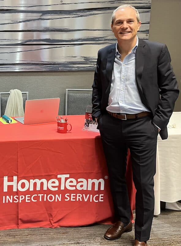 A smiling man in a dark suit and light blue shirt stands next to a table draped in a red tablecloth featuring the "HomeTeam Inspection Service" logo.