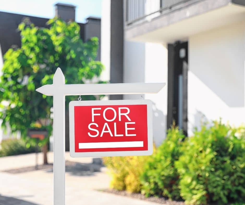 A red and white "For Sale" real estate sign hanging on a white wooden post in front of a modern residential house and garden.