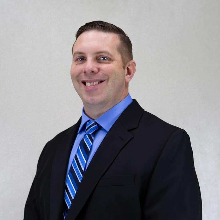 A professional portrait of a man with short brown hair smiling, wearing a black blazer, a light blue shirt, and a blue and white striped tie against a light grey background.