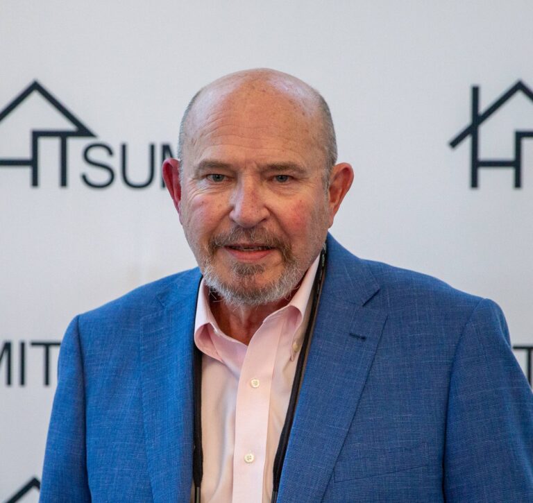 A man with a grey beard, bald head, and lanyard wearing a blue blazer over a light pink shirt in front of an HT Summit backdrop.