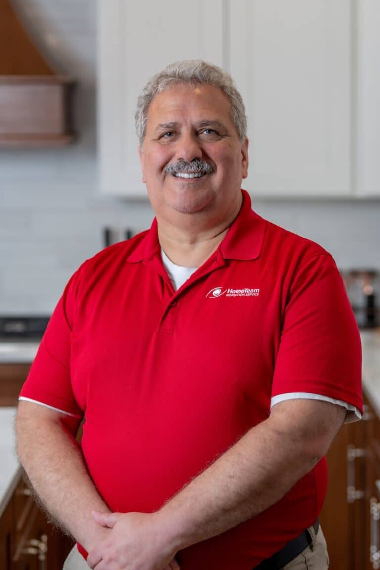 A smiling man with grey hair and a mustache wearing a red polo shirt, standing in a kitchen with his hands folded.