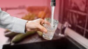 A hand holding a clear glass under a running kitchen faucet to fill it with tap water.